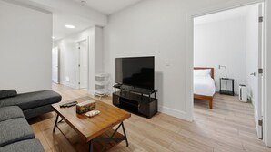 Ein modernes Hotelzimmer mit Flachbildfernseher, Esstisch aus Holz, einer Couch, einem Bett und einem kleinen Nachttisch.