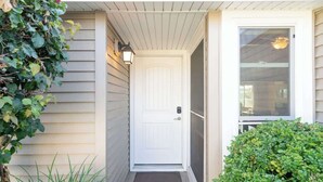 Uma porta branca com tapete, um pequeno alpendre com luminária e uma janela com vista para o interior.