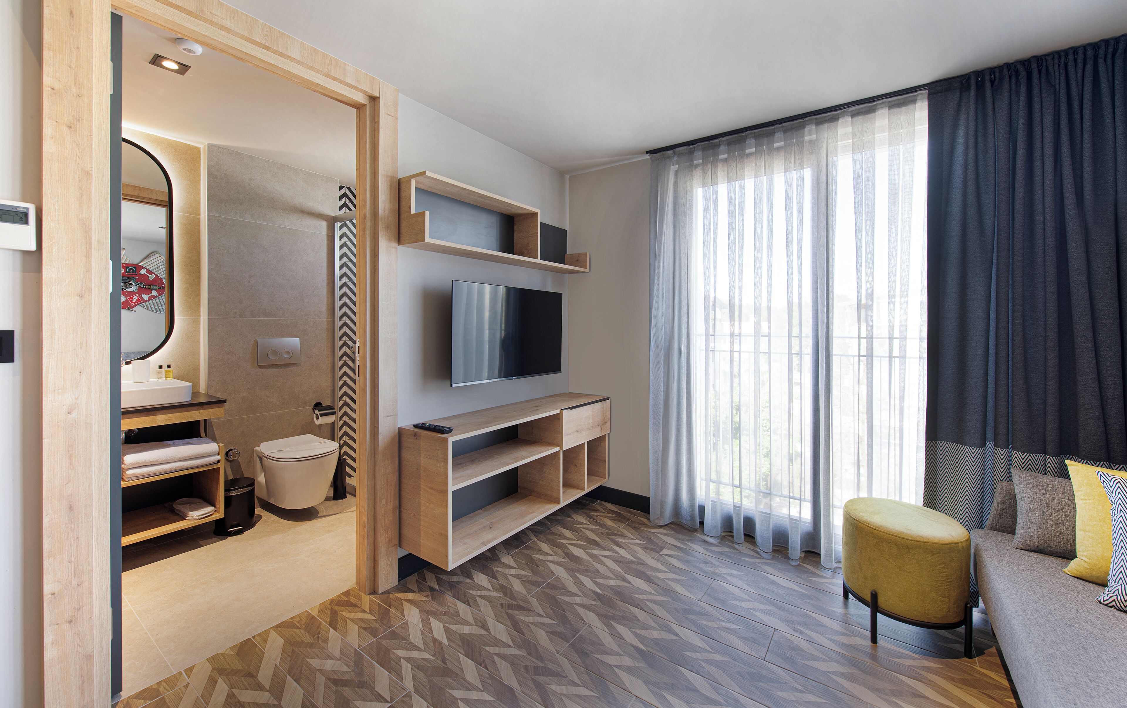 A modern hotel room with a wooden vanity, a flat-screen TV, a sofa, and a large window with curtains.