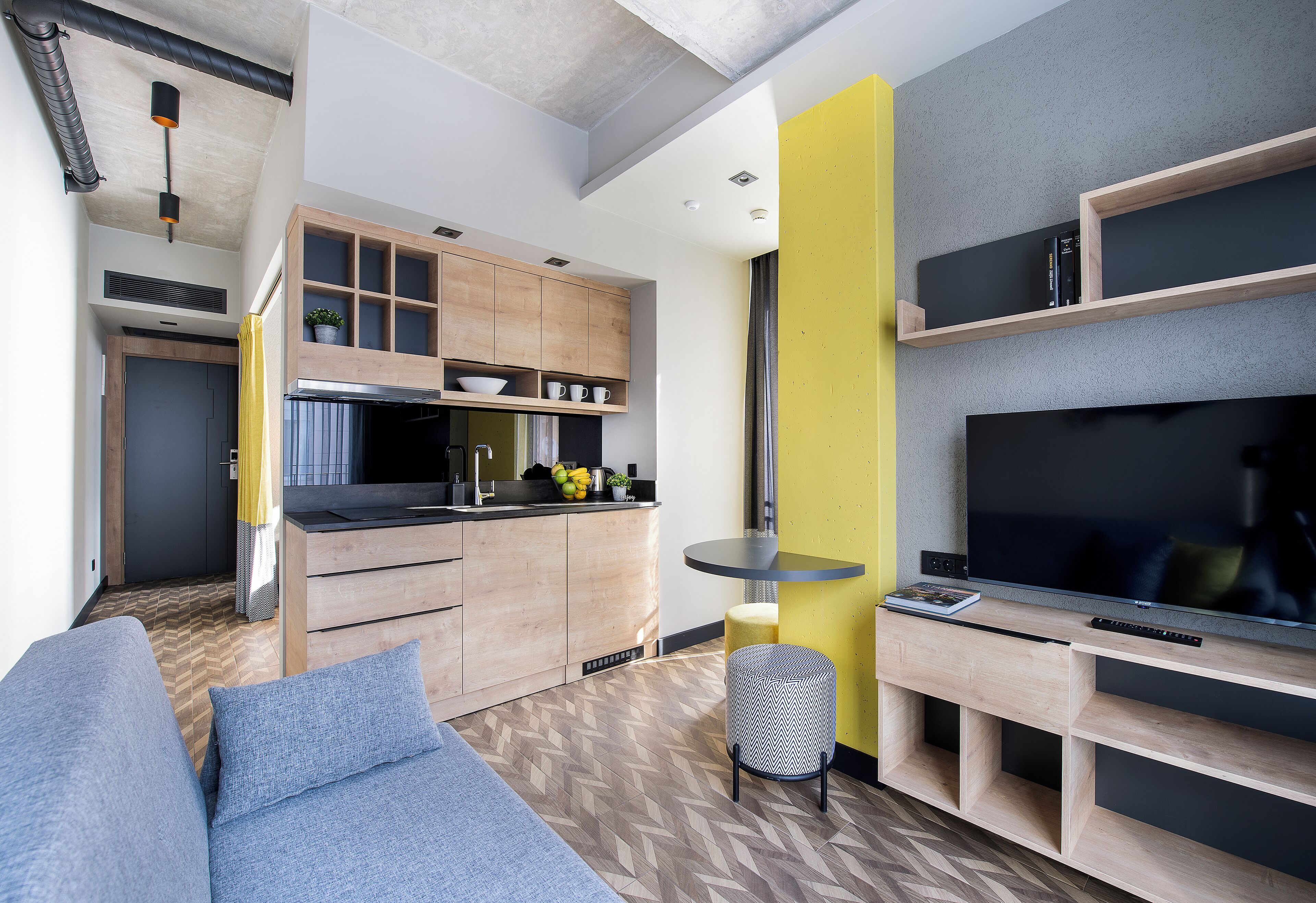 A modern living room with a sofa, a TV, and a small table. There is a kitchen area with cabinets and a countertop.