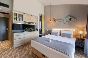 Una habitación de hotel moderna con una cama grande, una cocina integrada con armarios de madera, un original cuadro de peces y una mesita de noche pequeña.
