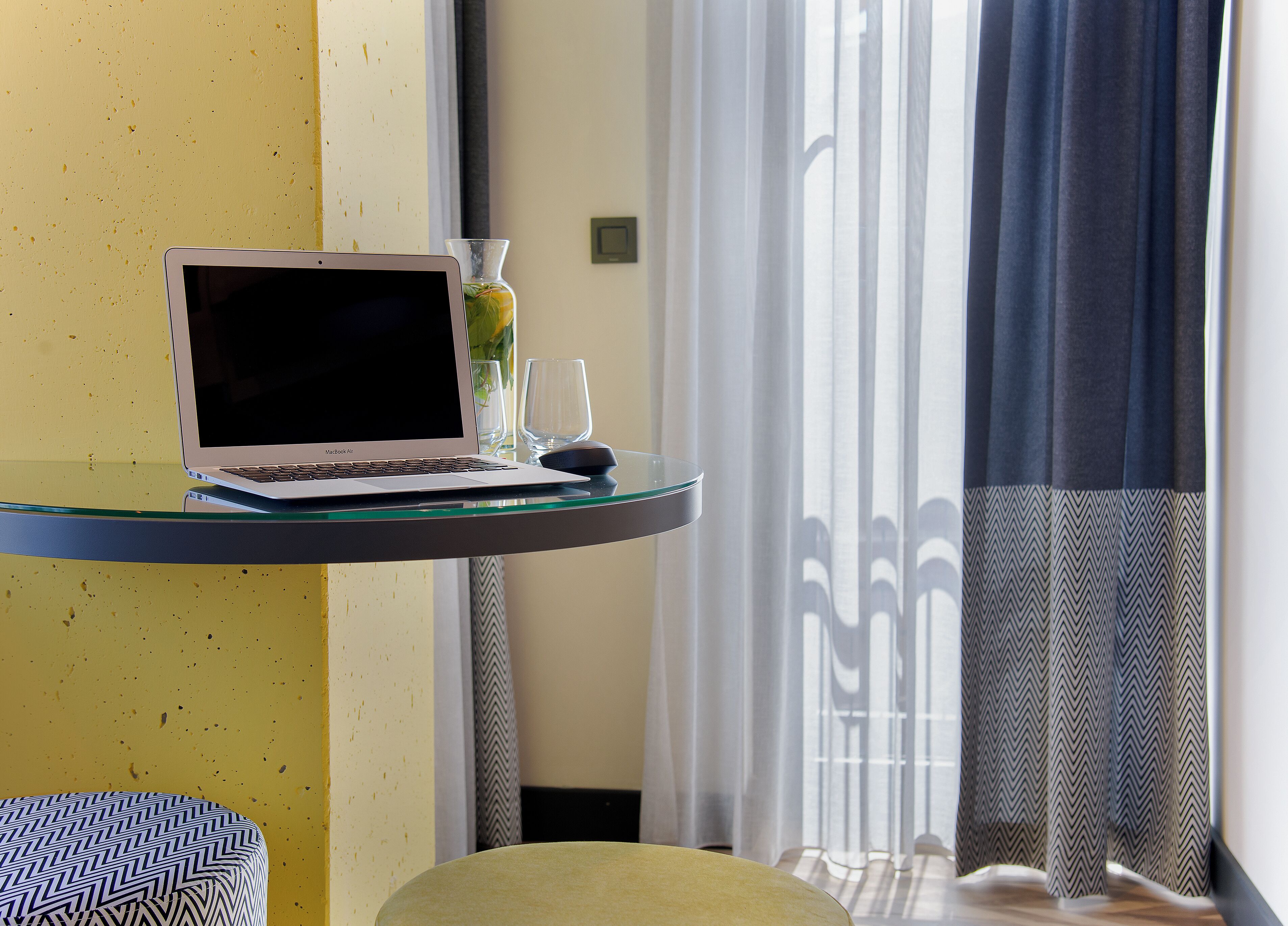A laptop on a glass table with a vase and a glass on it, a chair, and a window with curtains.