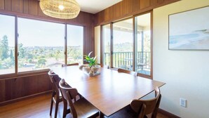 Uma área de refeições com uma grande mesa de madeira, cadeiras, luminária pendente e amplas janelas com vista para o exterior.