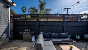 Ein Garten mit Holzzaun, Grill, grauem Sofa und Holztisch mit Pflanze.