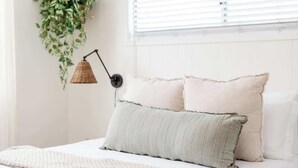Ein ordentlich bezogenes Bett mit zwei Kissen, eine hängende Pflanze und eine Lampe an einer weißen Wand.