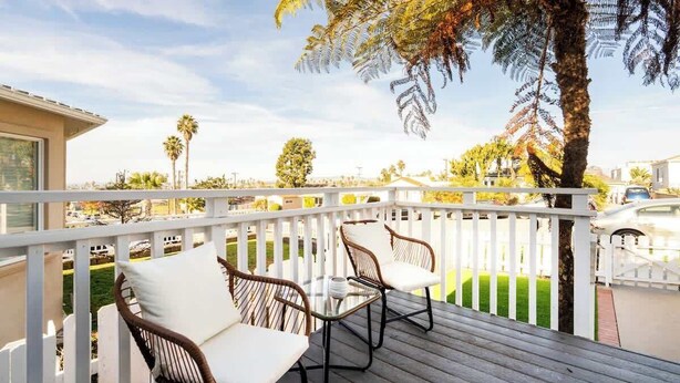 Une terrasse en bois, agrémentée de chaises en osier et d’une table en verre, qui offre une vue sur un quartier résidentiel avec des palmiers et des maisons.