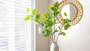 A round glass table with a checkered coaster, a white vase with green foliage, and a round mirror with a woven frame. - Sunny 3BR Seaside Escape | Beach & Bay Steps Away! (San Diego)