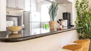 A kitchen with white cabinets, a black countertop, and a large plant. There are three wicker bar stools with white cushions. - Sunny 3BR Seaside Escape | Beach & Bay Steps Away! (San Diego)