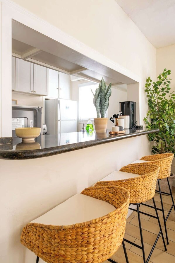 A kitchen with white cabinets, a black countertop, and a large plant. There are three wicker bar stools with white cushions.