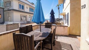 Um pátio externo com mesa e cadeiras de madeira, dois guarda-sóis azuis e vista para edifícios vizinhos.