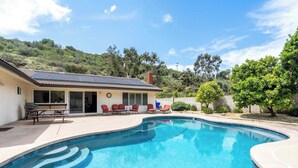 Une piscine à bords incurvés, une terrasse avec des chaises rouges et une maison dotée d’un toit équipé de panneaux solaires.