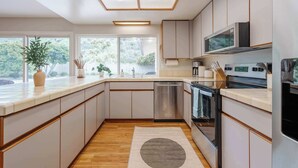 Une cuisine moderne avec des meubles blancs, un lave-vaisselle en acier inoxydable, un four à micro-ondes et un four traditionnel. Une grande fenêtre offre une lumière naturelle et une vue sur la verdure environnante.