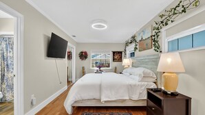 A bedroom with a bed, a nightstand with a lamp, a TV on the wall, and a window with a view. - Walk to Downtown Mount Dora! Patio Oasis + King Bed! (Mount Dora)