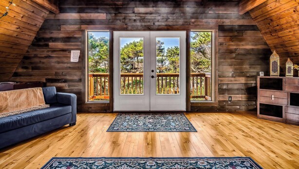 A cozy attic room with wooden walls, a blue sofa, and a large window with a view of the outdoors.