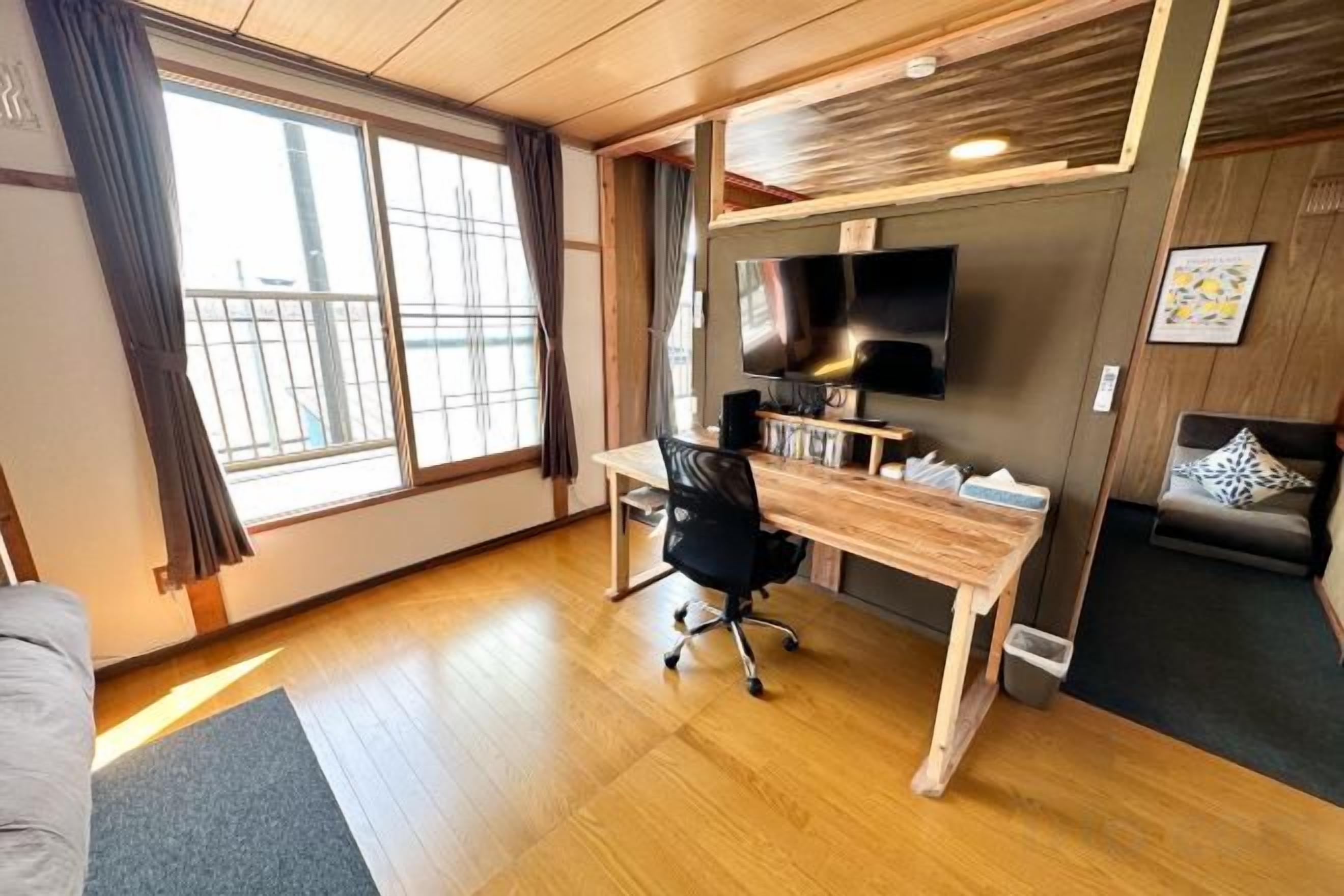 A room with a wooden desk, a black office chair, a television on the wall, and a sofa with a patterned cushion.