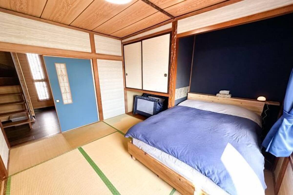 Et traditionelt japansk værelse med tatamimåtter, en seng med et blåt sengetæppe, en skydedør og et lille skrivebord med en lampe.