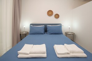 Una cama bien hecha con ropa de cama azul y toallas blancas, dos lámparas fijadas a la pared y una pequeña mesa redonda con una lámpara.