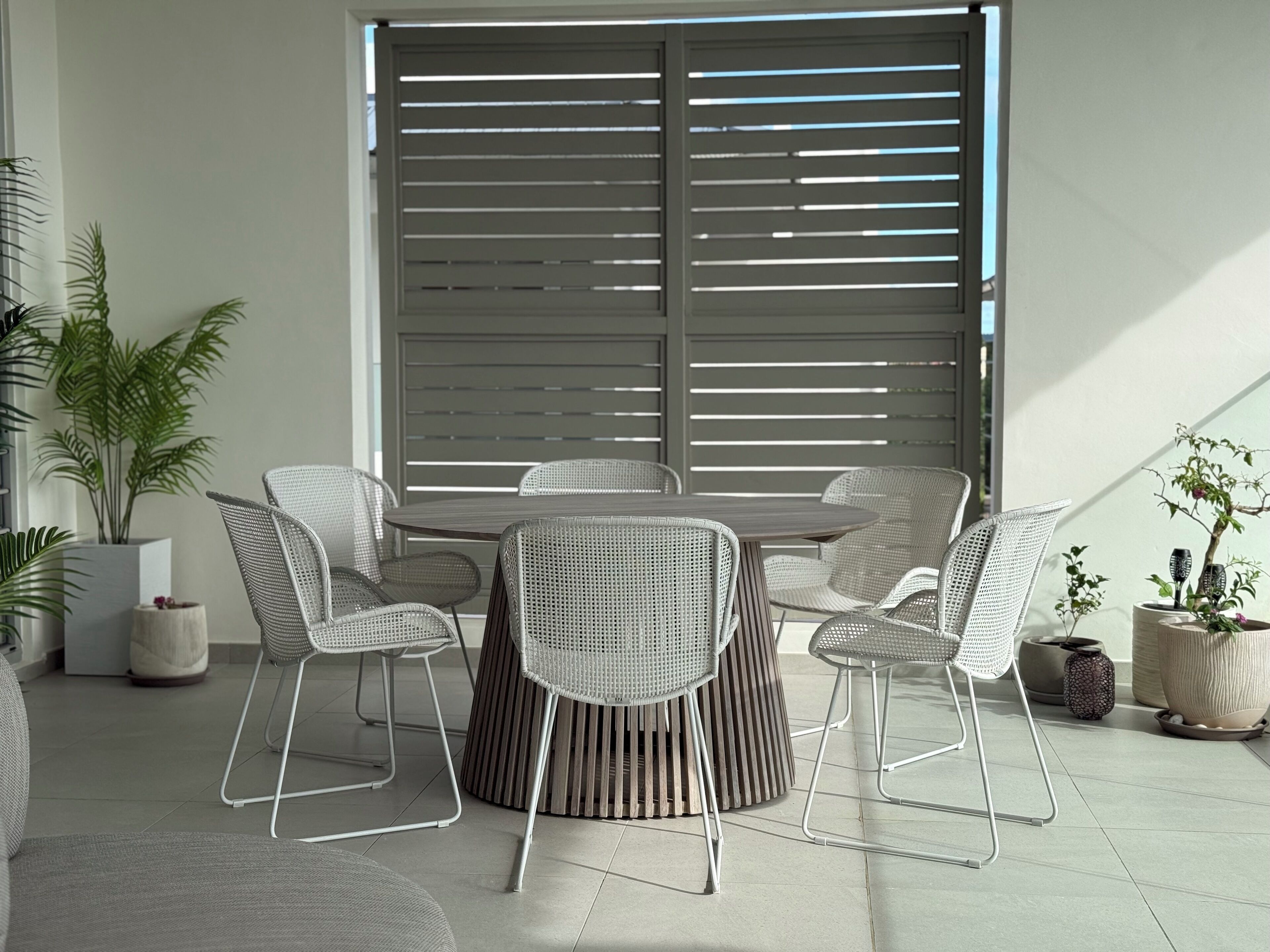 A dining area with a round table and six chairs, surrounded by potted plants and a large window with horizontal blinds.