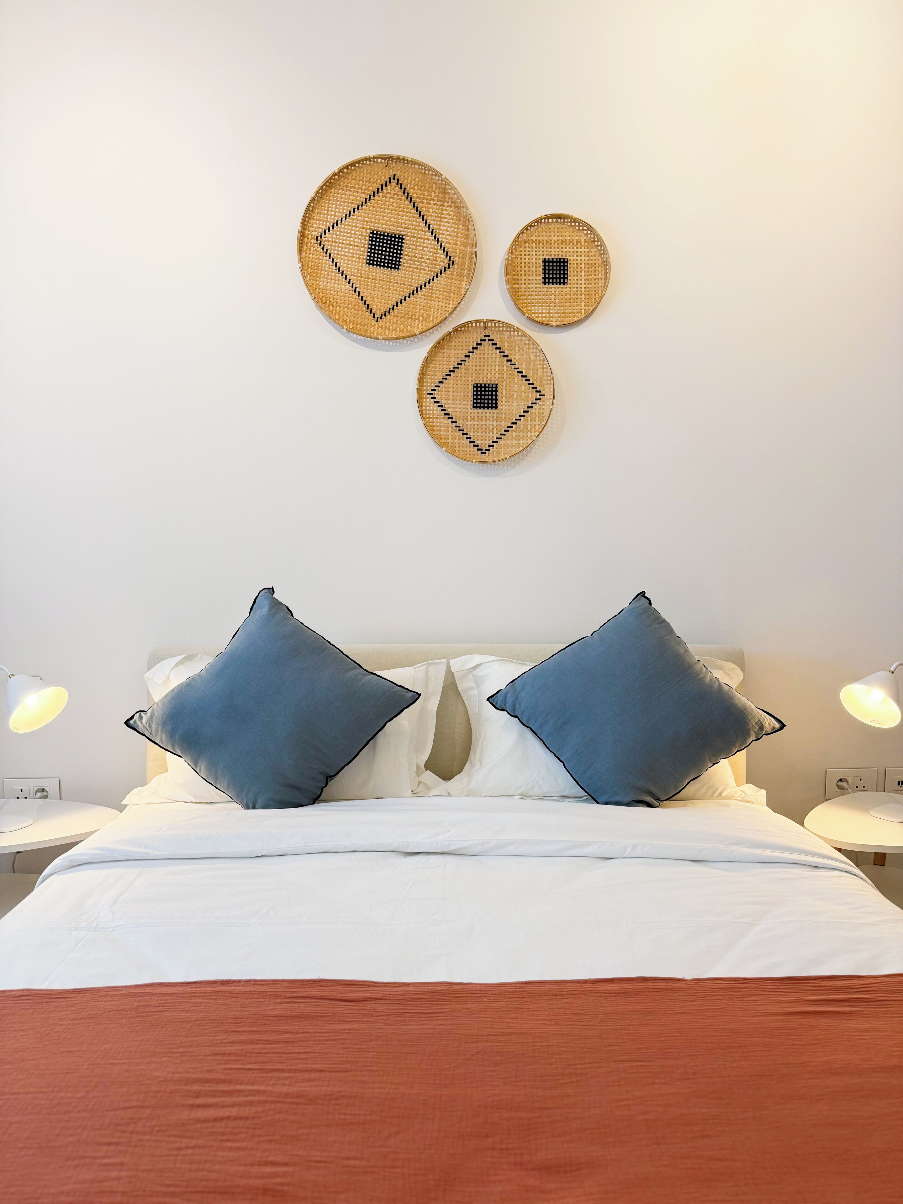 A neatly made bed with white bedding and two blue pillows, a bedside lamp, and three woven wall decorations.