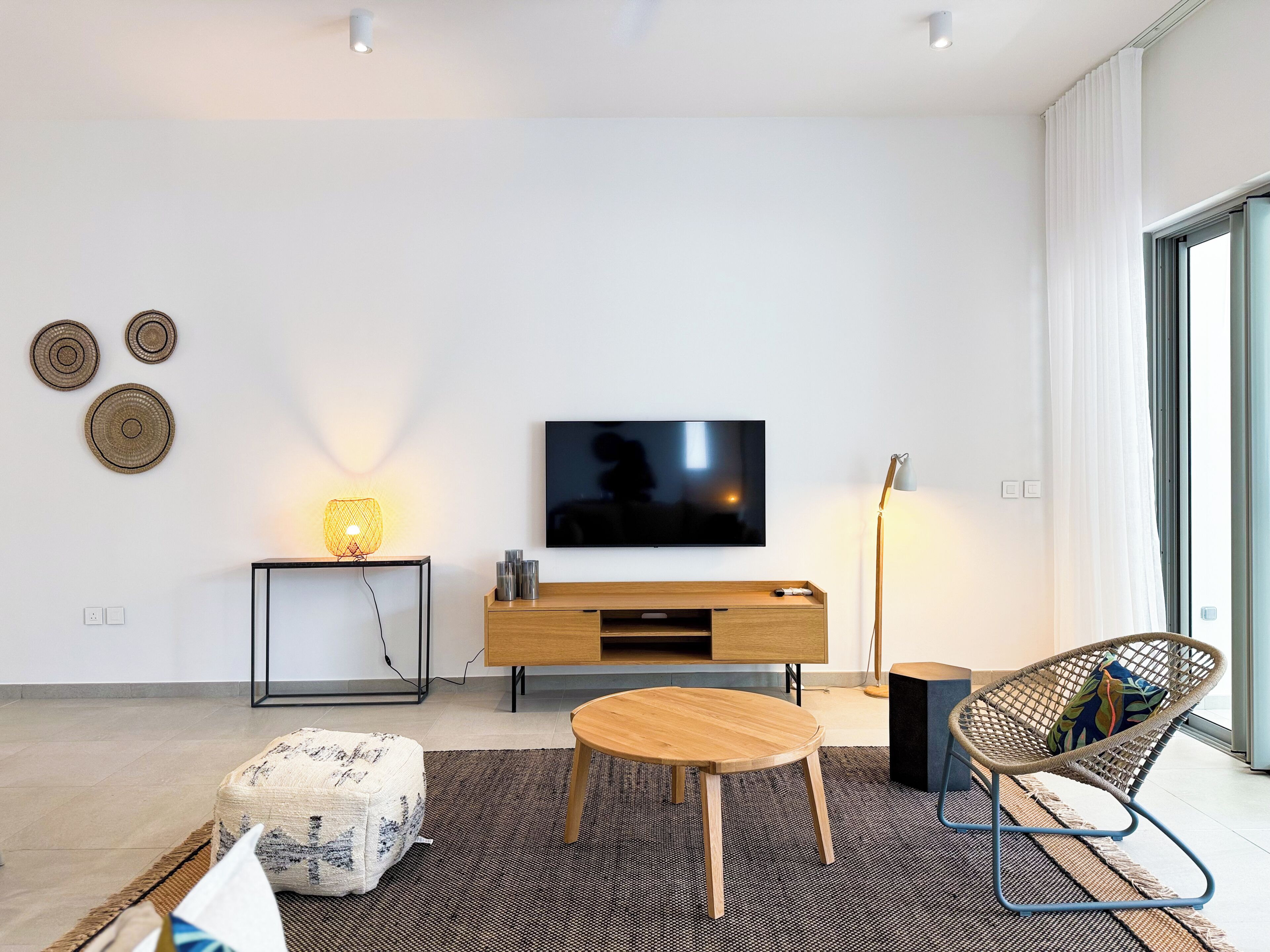 A modern living room with a flat-screen TV, wooden furniture, a round coffee table, and a large window.