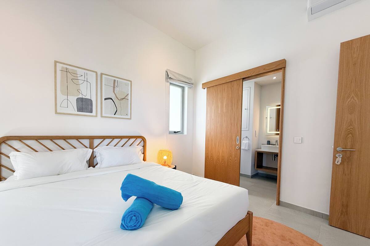 Una cama bien hecha con una toalla enrollada, una lámpara de noche y dos cuadros abstractos en la pared.