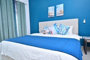 A neatly made bed with a blue and white bedspread, two framed pictures of seashells on the wall, and a green curtain.