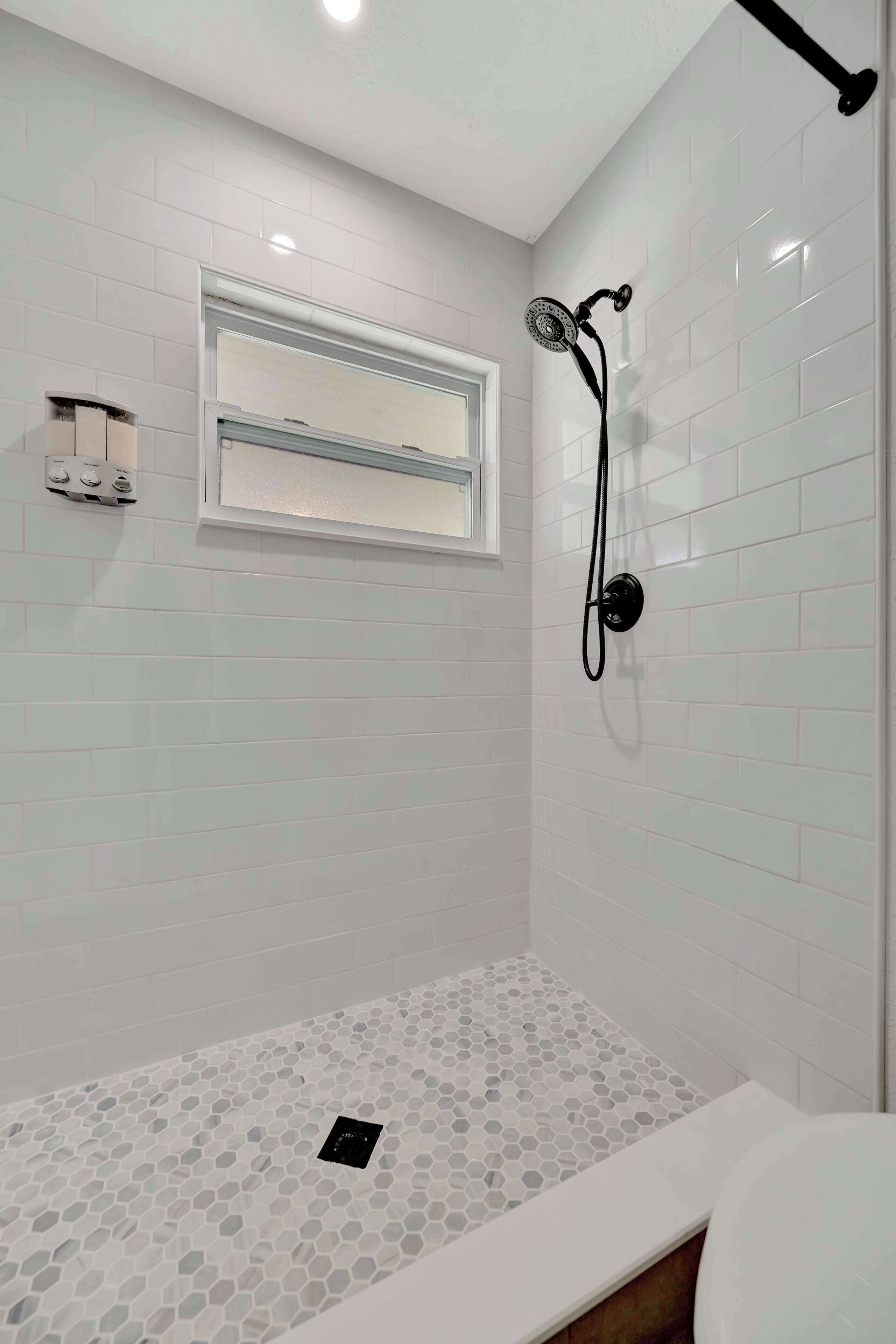 A bathroom with white tiled walls, a shower with a handheld showerhead, and a small window.