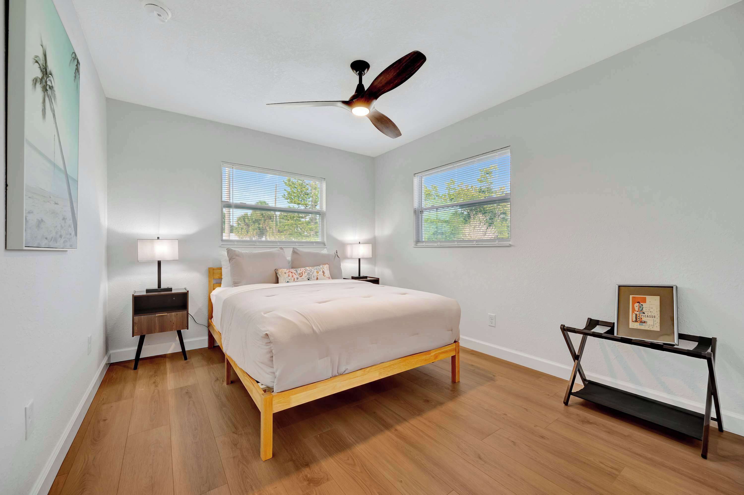 Un dormitorio con una cama, dos mesitas de noche, un ventilador de techo y dos ventanas.