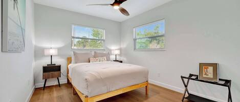 Un dormitorio con una cama, dos mesitas de noche, un ventilador de techo y dos ventanas.