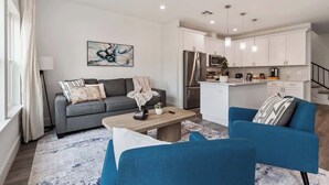 Ein modernes Wohnzimmer mit einem grauen Sofa, blauen Sesseln und einem Holztisch. Im Hintergrund ist eine Küche mit weißen Schränken und Edelstahlgeräten zu sehen.