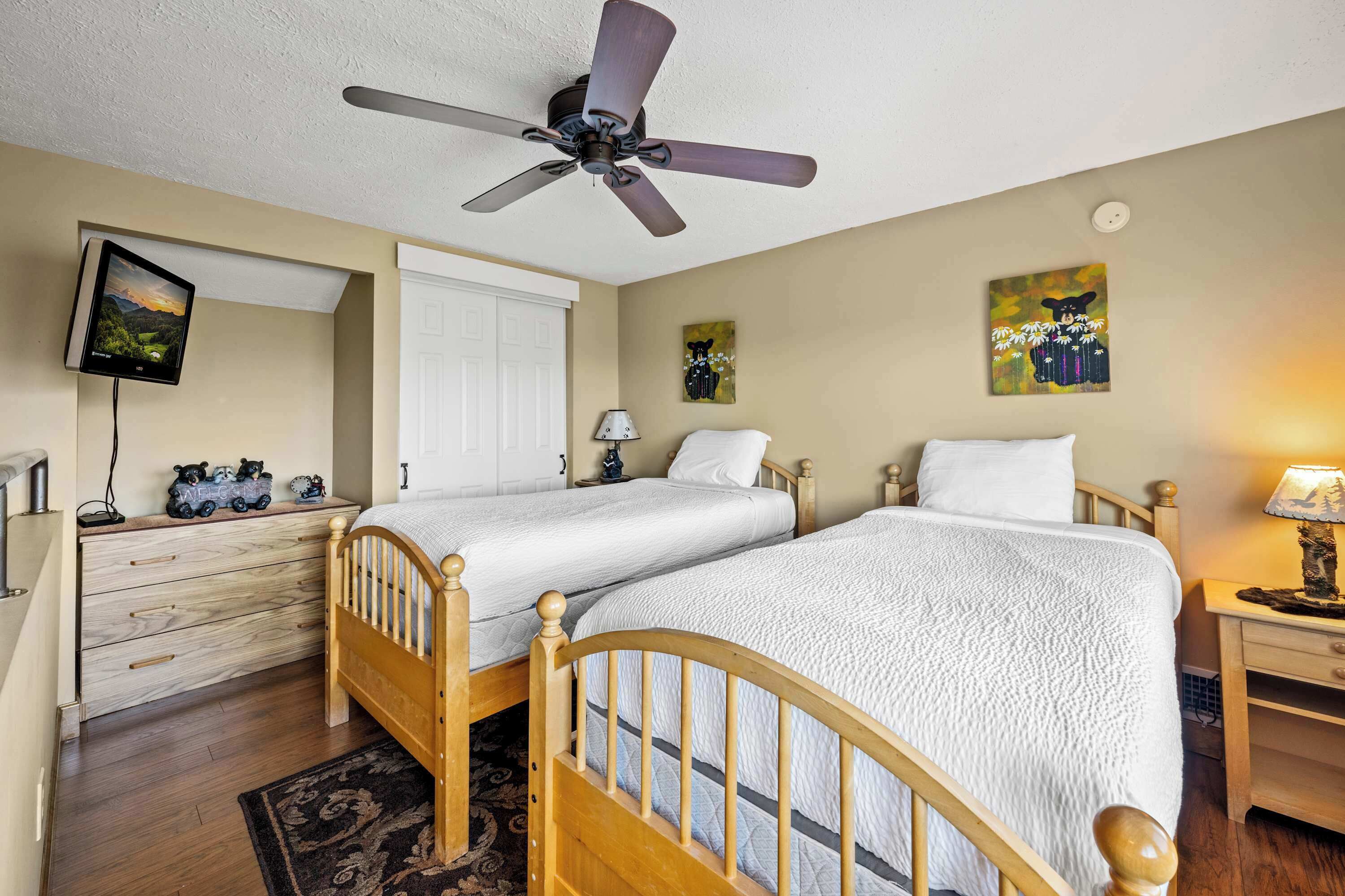 A bedroom with two wooden beds, a ceiling fan, a TV mounted on the wall, and artwork on the walls.