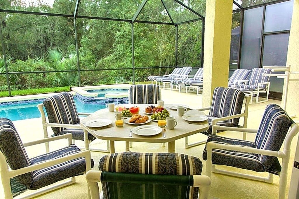 A covered outdoor seating area with a pool, chairs, and a table set for breakfast.