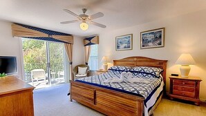 A bedroom with a wooden bed, a ceiling fan, a TV, a desk, and a balcony with chairs.