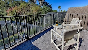A balcony with a view of a road and trees, a small table with a bottle, and two striped chairs.