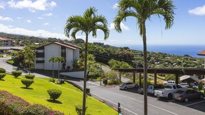 A coastal road with parked cars, palm trees, and a building with a white facade. - Kona Coffee Villas E246 2 Bedroom Condo (Holualoa)