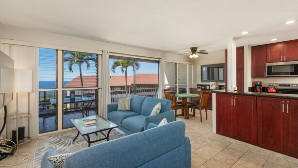 A spacious living area with a blue sofa, a dining table, and a kitchen with wooden cabinets and a microwave. - Kona Coffee Villas E246 2 Bedroom Condo (Holualoa)