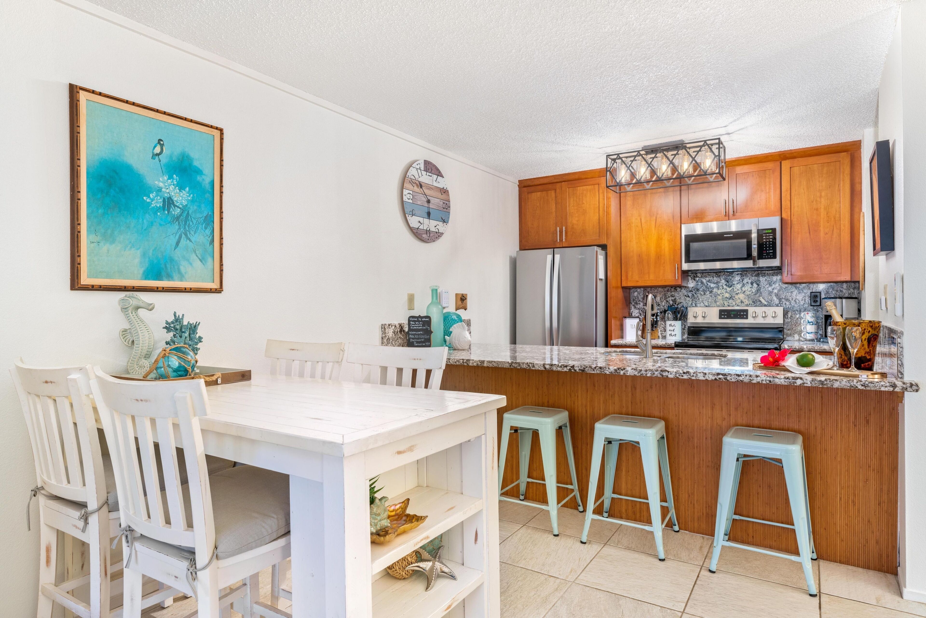 A kitchen with wooden cabinets, a stainless steel refrigerator, a microwave, and a granite countertop. There are two light blue barstools and a white dining table with four chairs.