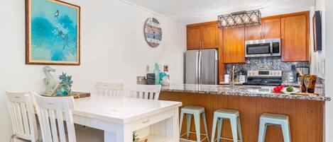 A kitchen with wooden cabinets, a stainless steel refrigerator, a microwave, and a granite countertop. There are two light blue barstools and a white dining table with four chairs.
