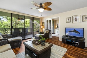 A modern living room with wicker furniture, a ceiling fan, a flat-screen TV, and large windows offering a view of the outdoors. - Kona Pacific D526 1 Bedroom Condo (Kailua-Kona)