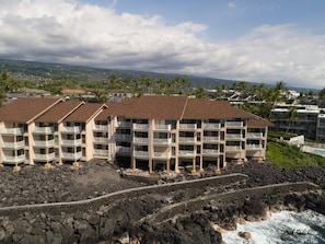 A beachfront hotel with multiple balconies, a rocky shoreline, and a view of the ocean and distant landscape. - Sea Village 4313 2 Bedroom Condo (Kailua-Kona)