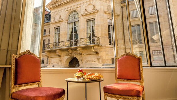 Une pièce comprenant deux fauteuils rouges à revêtement en tissu et une petite table ronde sur laquelle se trouvent un panier de fruits et des pâtisseries. Une grande fenêtre offre une vue sur un bâtiment historique à l’architecture riche et ornée.