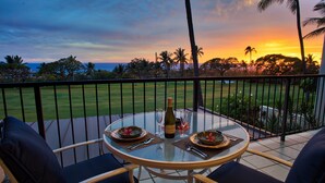 Une table ronde en verre, accompagnée de deux chaises, est placée sur un balcon qui offre une vue sur un terrain de golf et des palmiers, le tout baigné par les couleurs du coucher de soleil.