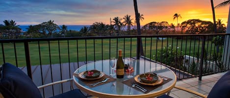 A round glass table with two chairs on a balcony overlooking a golf course and palm trees during sunset.