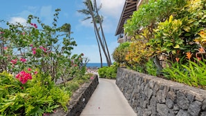 A pathway lined with lush greenery and vibrant flowers, leading towards a building with a stone wall and a view of the ocean. - Kona Makai 3305 2 Bedroom Condo (Kailua-Kona)