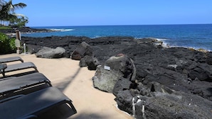 A beachside view with a rocky shoreline, lounge chairs, and a clear blue sky. - Kona Makai 3305 2 Bedroom Condo (Kailua-Kona)