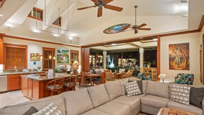A spacious living area with a large sofa, dining table, and chairs, a kitchen area with wooden cabinets, and a ceiling fan.