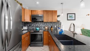 A kitchen with wooden cabinets, a stainless steel refrigerator, a built-in oven, a microwave, a granite countertop, a black sink, and a backsplash with a mosaic tile pattern.