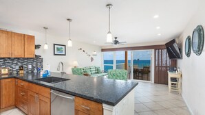 A modern kitchen with wooden cabinets, a granite countertop, and a built-in dishwasher. There is a dining area with a green and white checkered sofa and a view of the ocean through large sliding glass doors.