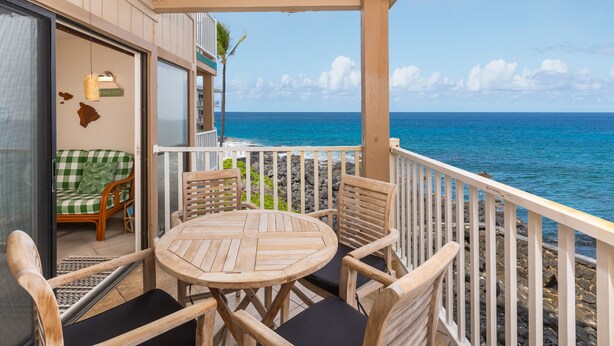 A wooden deck with a round table and chairs, a green and white checkered sofa, and a view of the ocean.