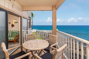 A wooden deck with a round table and chairs, a green and white checkered sofa, and a view of the ocean. - Sea Village 4113 2 Bedroom Condo (Kailua-Kona)